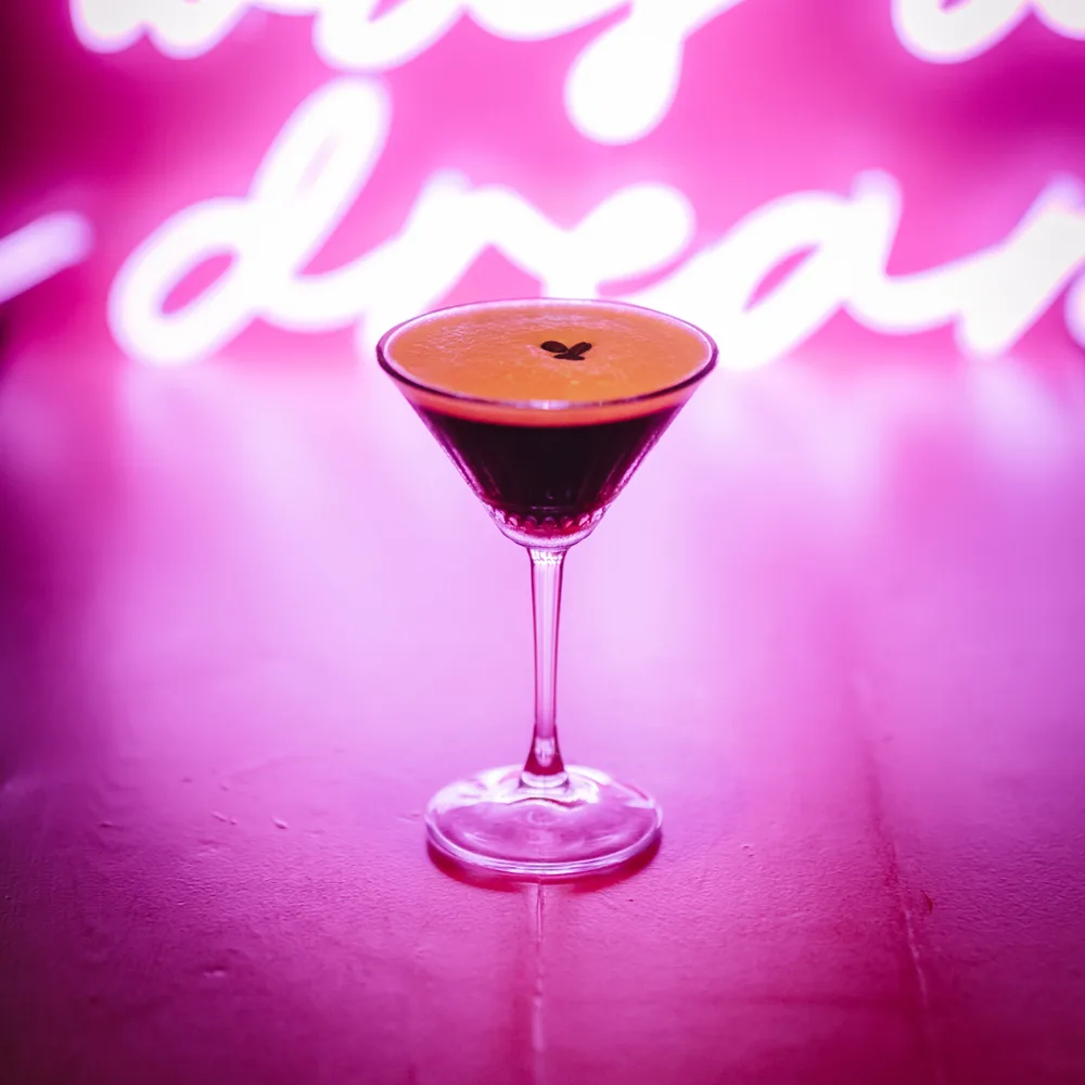 Donkere cocktail in een coupeglas voor een roze neonbord.