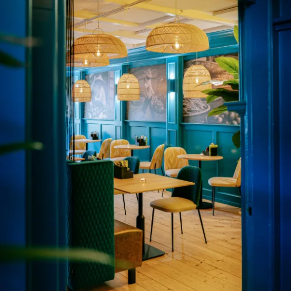 Doorkijk in de eetzaal met blauwe lambrisering, rotan lampen en okergele stoelen.