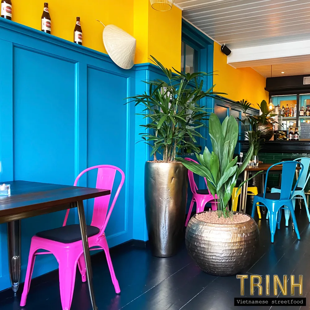 Interieur met knalblauwe lambrisering, gele muur, roze en blauwe stoelen en planten.