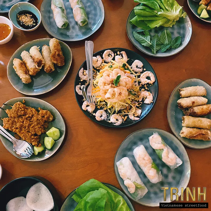 Grote gedekte tafel met garnalensalade, loempia's, gỏi cuốn en schaaltjes verse kruiden.