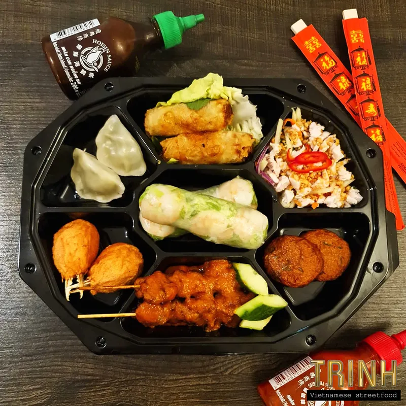 Zwarte bentobox met dumplings, loempia's, saté en salade, met chilisaus en eetstokjes ernaast.