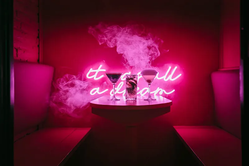 Roze neonbord met de tekst "it was all a dream" boven een tafel met twee cocktails.