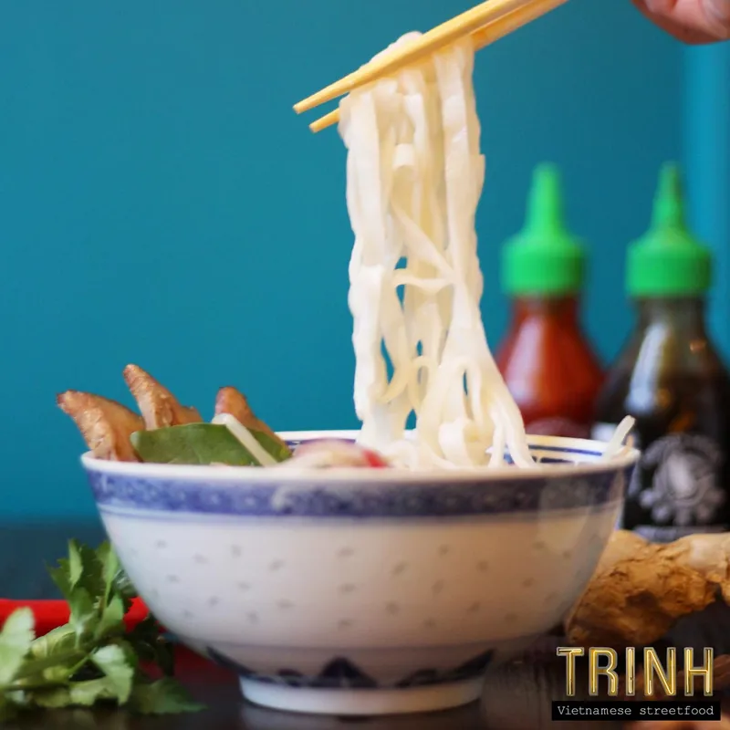 Eetstokjes tillen rijstnoedels uit een kom phở, met sausflessen op de achtergrond.