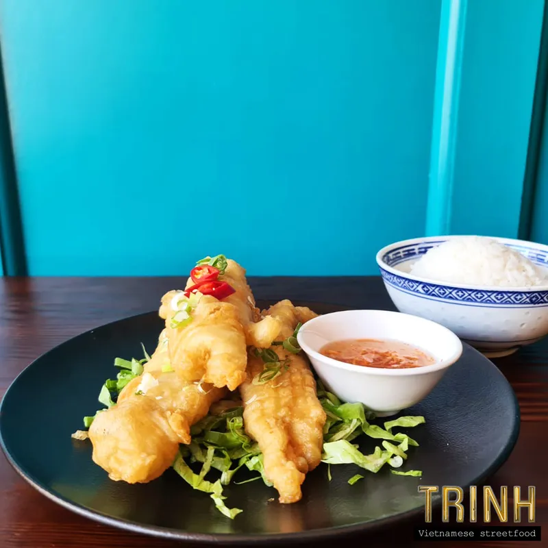 Bord gefrituurde vis met chilisaus en een kommetje rijst tegen een turquoise achtergrond.