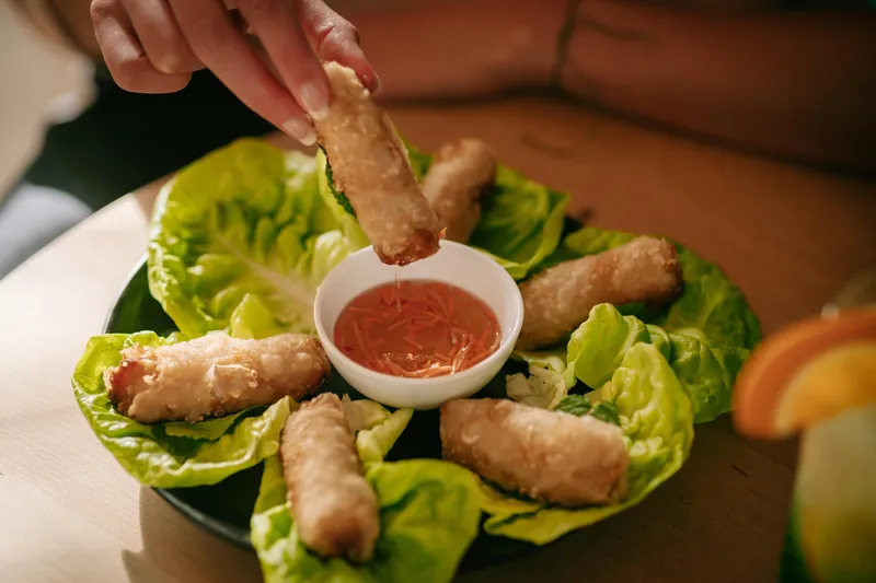 Hand doopt een loempia in zoetzure saus, omringd door loempia's op slablaadjes.