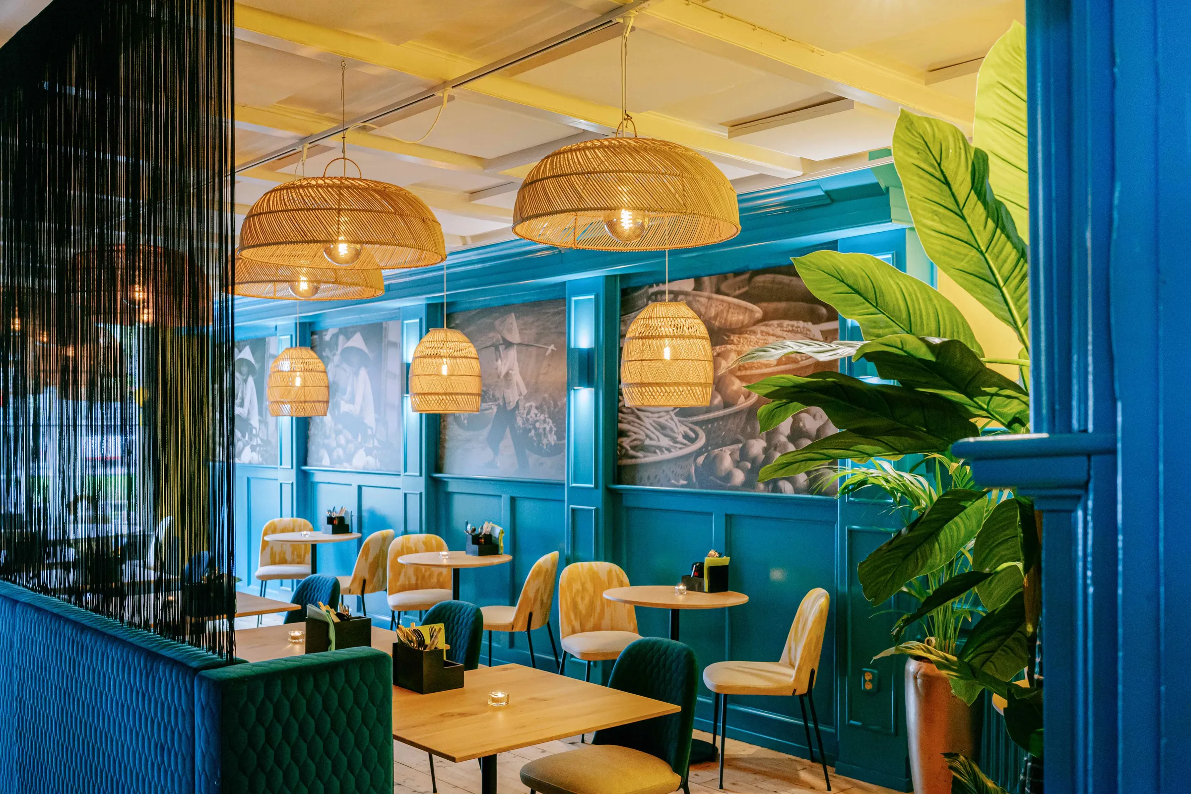 Eetzaal van het restaurant met turquoise lambrisering, rotan hanglampen, groene banken en planten.