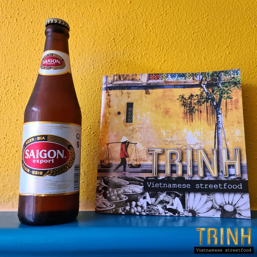 Flesje Saigon Export bier naast het menuboekje van Trinh tegen een gele muur.