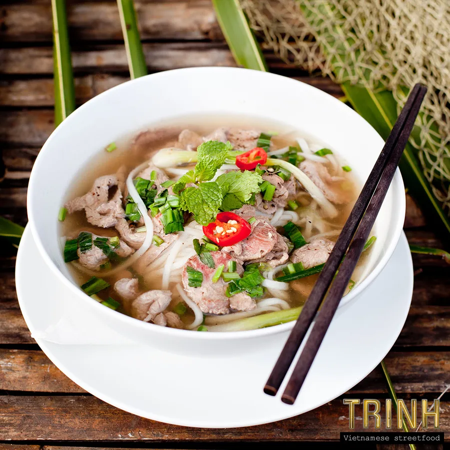 Kom phở bò met rundvlees, munt en rode peper, met eetstokjes op een bamboe ondergrond.